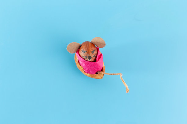 可爱的老鼠手工艺品，躺在粉色布料制成的坚果床上，背景为浅蓝色，展现出一个奇幻而温馨的秋季主题场景。图片下载