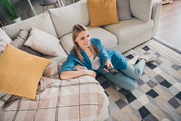 俯视照片：年轻的居家女孩坐在现代设计公寓的客厅地毯上，使用遥控器观看电视。图片下载