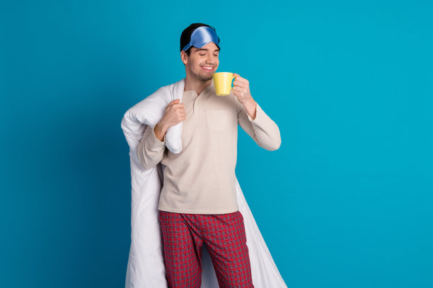 漂亮的年轻男性的照片肖像拿着羽绒被闻咖啡穿睡衣过夜在蓝色的背景隔离晚安图片下载