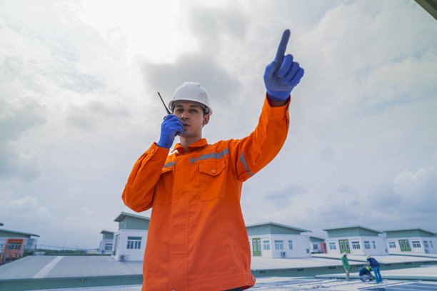 穿着橙色安全夹克的男性工程师，在监督建筑屋顶太阳能电池板的安装时进行沟通。可持续技术解决方案的太阳能项目。图片下载