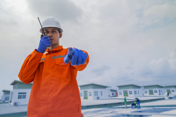 穿着橙色安全夹克的男性工程师，在监督建筑屋顶太阳能电池板的安装时进行沟通。这是可持续技术解决方案的太阳能项目。图片下载