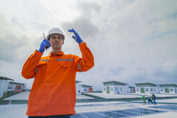 穿着橙色安全夹克的男性工程师在监督建筑屋顶太阳能电池板的安装时进行沟通。可持续技术解决方案的太阳能项目。图片下载
