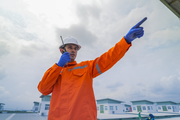 一名工程师，穿着橙色安全夹克，在监督建筑屋顶太阳能电池板的安装时进行沟通。这是可持续技术解决方案的太阳能项目。图片下载