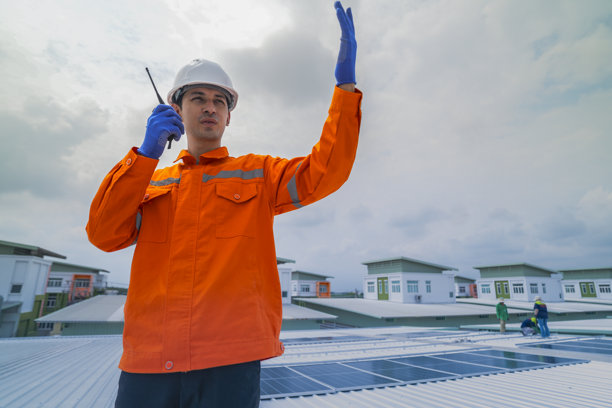 穿着橙色安全夹克的男性工程师，在监督建筑屋顶太阳能电池板的安装时进行沟通。这是可持续技术解决方案的太阳能项目。图片下载