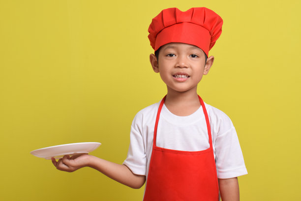 可爱的小厨师手拿盘子。一个穿着红色围裙和帽子的亚洲男孩。图片下载