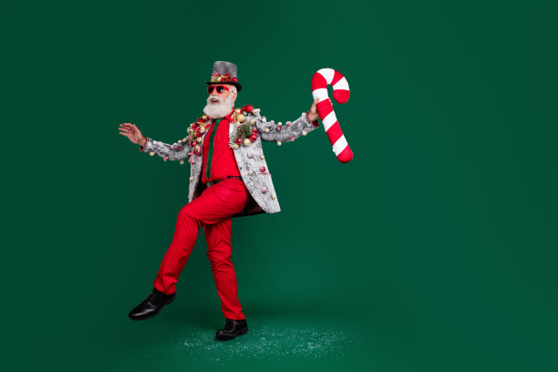 全身照，孤立的兴奋男人穿着红色裤子，身旁有圣诞树、玩具，穿着西装外套，手持糖果棒，背景为深绿色，留有空白空间。图片下载