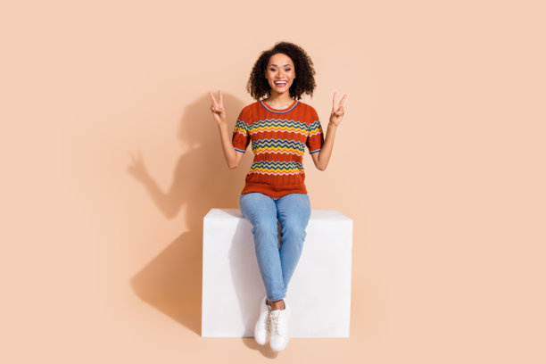 一个穿着针织t恤牛仔裤的快乐女人的全尺寸照片，坐在立方体上，在米色背景上显示v字符号图片下载