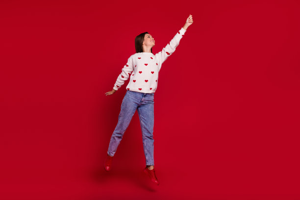 穿着白色毛衣的可爱女孩在红色背景下高高跳起，伸手抓星星的全身照片图片下载