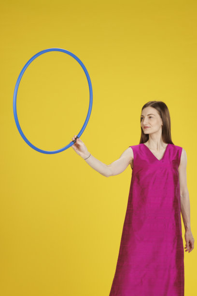 充满活力的肖像，漂亮的女商人正在练习呼啦圈。紫红色丝绸裙子，优雅的生活方式。黄色背景。图片下载