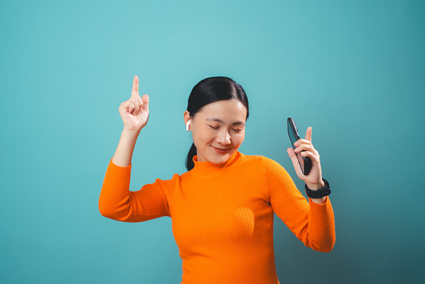 亚洲女性享受使用智能手机，戴着耳机听音乐，背景为单色。图片下载