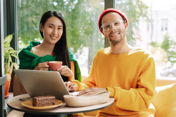 年轻快乐、微笑的情侣在咖啡馆使用笔记本电脑并喝咖啡，朝着镜头看。图片下载