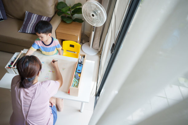 亚洲母亲正在桌子上与一个男孩用彩色铅笔画画。男孩坐在一把黄色的椅子上。图片下载