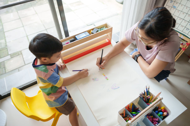 亚洲母亲和小男孩坐在小桌子旁，用彩色铅笔在客厅里涂色，周围是艺术用品。女人正在帮助孩子进行绘画。图片下载