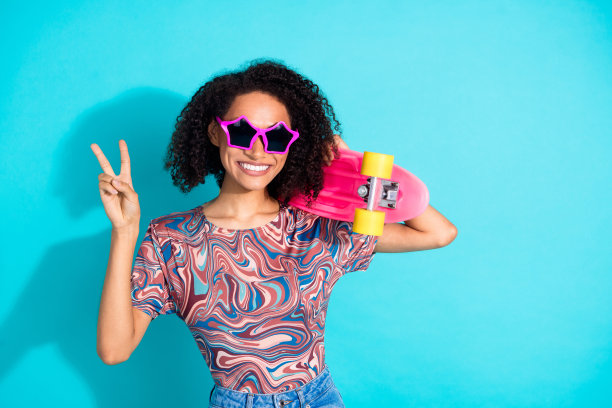 时尚年轻女孩的照片肖像，戴着太阳镜，手持滑板，做出V字手势，穿着色彩鲜艳的衣服，背景为水蓝色。图片下载