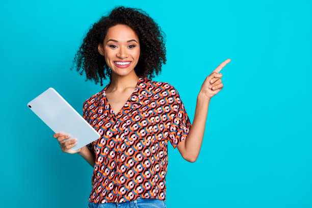 一位快乐的女孩，卷发披肩，穿着印花衬衫，手持平板电脑，指向空白的优惠空间，背景为蓝色。图片下载