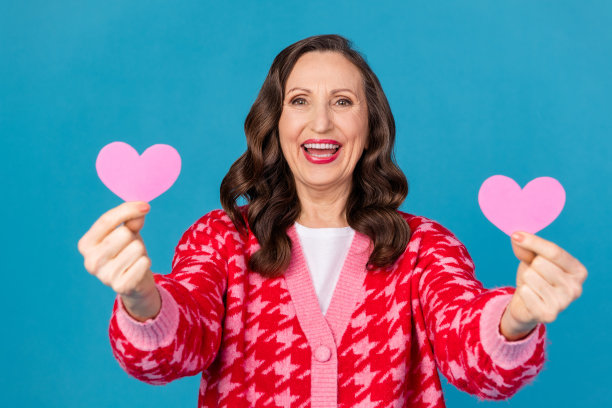 吸引人的退休女性的照片肖像，手持两个粉色心形物品，穿着时尚的粉色印花衣服，背景为蓝色。图片下载