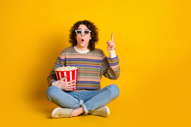 全身照，穿着毛衣的搞笑男士戴着3D眼镜，手持爆米花，举起手指，背景为黄色。图片下载