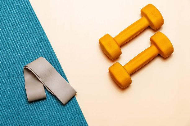 一对黄色哑铃放在桃色背景上。瑜伽垫上有一条弹力带。图片下载