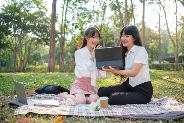 两个年轻女子坐在公园里的野餐毯上，享受着户外愉快的一天。图片下载