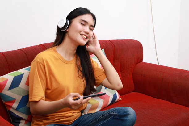 快乐的亚洲女性坐在沙发上听手机音乐，微笑的女孩在早晨放松，戴着耳机，享受放松时光。留白空间。图片下载