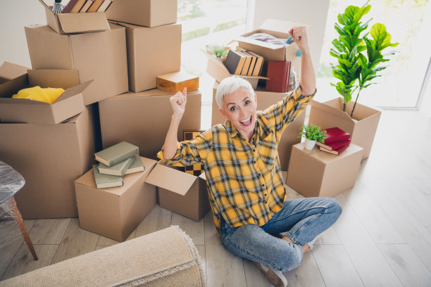 全景高角度照片，展现一位和蔼的老年女性坐在地板上，举起双拳，穿着格子衬衫，正在搬家到新公寓室内。图片下载