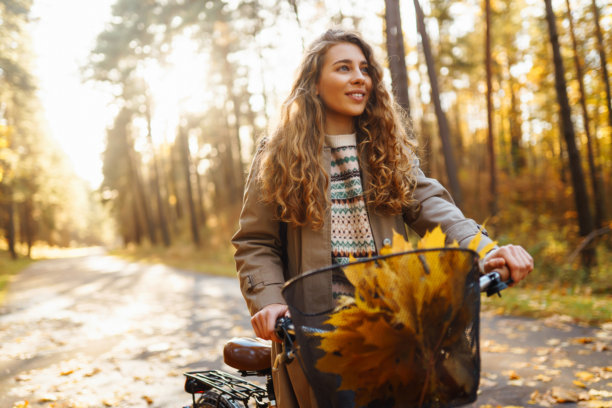 一位快乐的女性骑着自行车沿着风景如画的森林小径，阳光明媚的下午，地面上铺满了秋天的落叶。图片下载