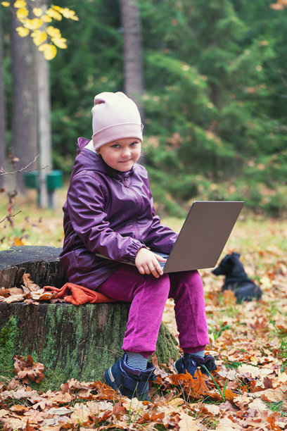 一个可爱的小女孩坐在森林中间的一根老树桩上，使用笔记本电脑进行在线学习或玩游戏。图片下载