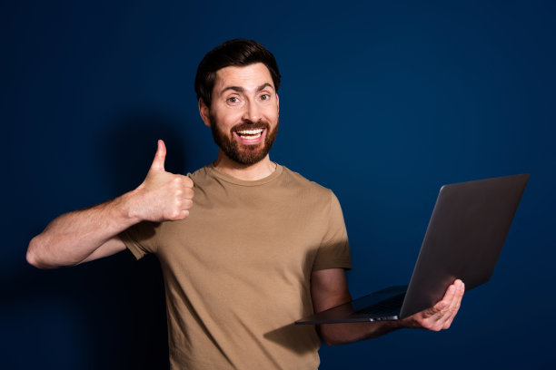 深色背景下，穿着米色T恤的棕发软件工程师男士在使用笔记本电脑工作，做出“点赞”手势的照片肖像。图片下载