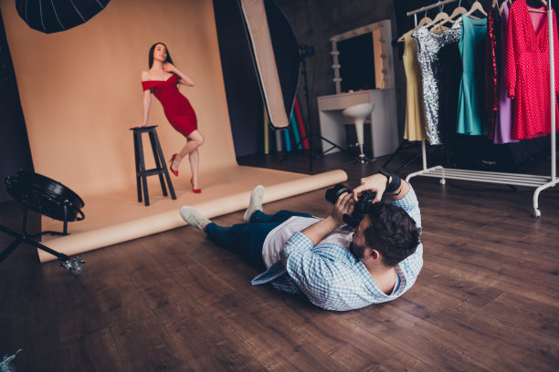 时尚男性摄影师躺着拍摄美丽模特在现代室内摄影棚的摆姿照片图片下载