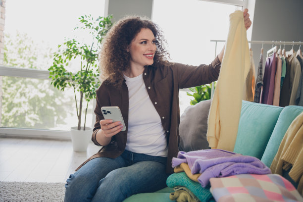 拿着智能手机的年轻有趣的女人在阁楼现代住宅内出售她的二手衣服，坐在沙发上的照片图片下载