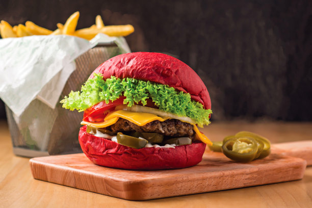 红色牛肉汉堡配生菜、墨西哥辣椒、西红柿和奶酪，放在木板上，旁边有薯条篮，快餐的侧面视图图片下载