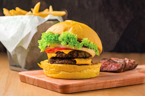 双层牛肉芝士汉堡，配生菜、墨西哥辣椒、西红柿、芝士和牛排片，放在木板上，旁边有一篮薯条，快餐的侧面视图图片下载