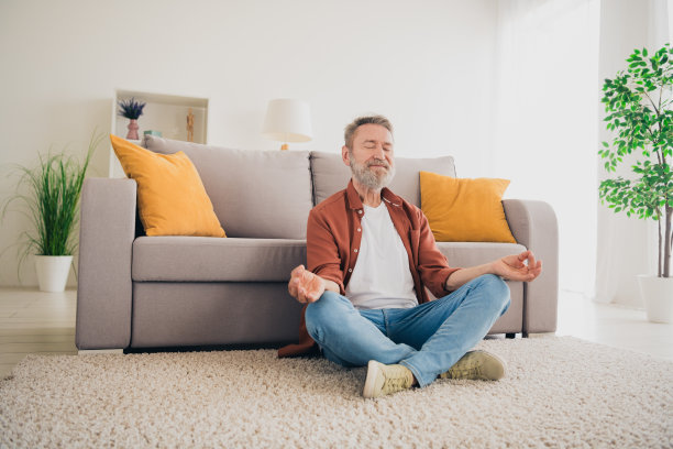 全身照片，和蔼的退休男士坐在地板上冥想，穿着棕色衣服，现代舒适的室内公寓环境。图片下载