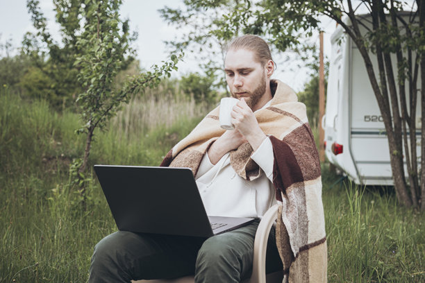 年轻的胡子露营者在户外享受休闲时光，手里拿着笔记本电脑和一杯咖啡。图片下载