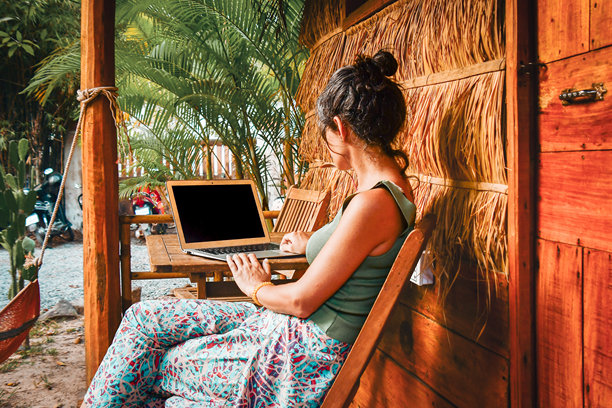 背影的白人嬉皮士女性坐在电脑旁，位于环保的稻草房建筑的客栈外。远程在东南亚的自由职业工作，孤立于热带自然环境中。图片下载