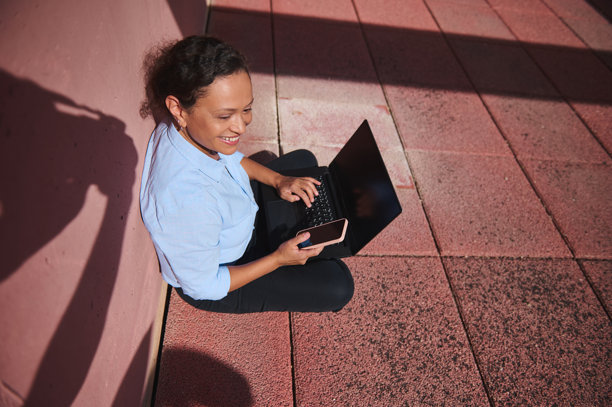 微笑的女性坐在户外，使用笔记本电脑和手机，在阳光明媚的日子里享受远程工作，坐在粉色瓷砖表面上。图片下载