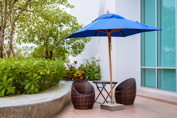 户外座位区的照片，配有蓝色伞、两把藤椅和一张小桌子，周围环绕着绿色树木。图片下载