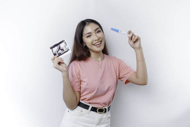 快乐的年轻女性穿着粉色T恤，展示她的怀孕测试和超声波照片。图片下载