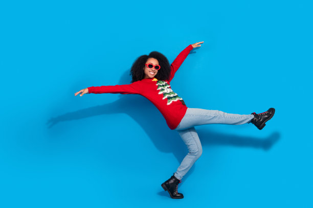 穿着红色圣诞服装的可爱年轻女士在蓝色背景下戴着太阳镜跳舞，庆祝冬季假期的全身照片图片下载