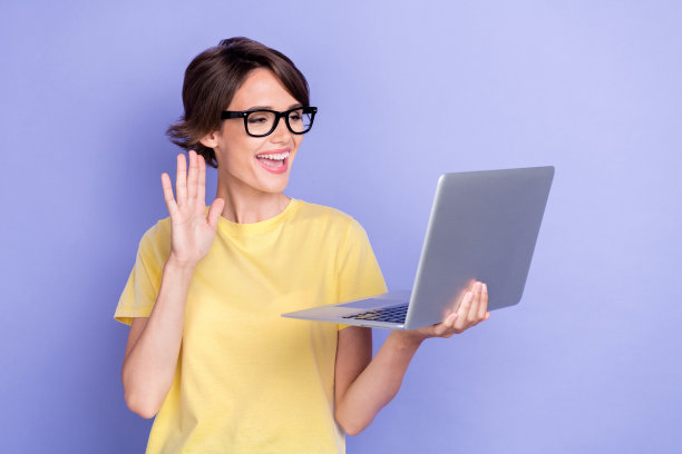 年轻迷人的可爱女性穿着黄色T恤，戴着眼镜，手持电脑，微笑着打招呼，背景为紫色。图片下载