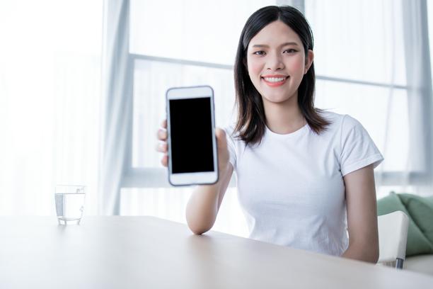 一位年轻女性坐在桌子旁，手里拿着一部智能手机。图片下载