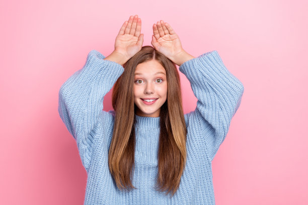 特写照片：有趣的女孩学生穿着蓝色针织毛衣，手里拿着兔子符号，头上玩耍，背景为粉色。图片下载