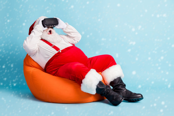 全身照片，退休老人留着胡子，惊讶地看到一只坐在橙色豆袋上的动画真实恐龙，穿着圣诞老人服装，系着吊带，戴着太阳镜和靴子，背景为蓝色。图片下载