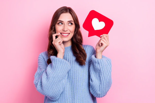 特写照片：穿着蓝色针织毛衣的女士，咬着指甲，带着梦幻般的表情，手持“喜欢”符号，背景为粉色。图片下载