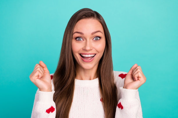 穿着白色套头衫的幸运女孩笑着举起拳头，隔离在蓝绿色背景上的照片图片下载