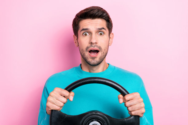 年轻男子的照片肖像，手握方向盘，表现出惊讶的反应，穿着时尚的蓝色衣服，背景为粉色。图片下载