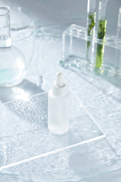 这则极简主义护肤广告展示了一瓶精华液放在带水滴的玻璃盘上，水面荡漾着涟漪，旁边还有装有化学物质和天然成分的玻璃实验室烧瓶。图片下载