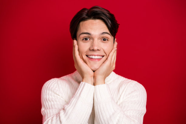 特写照片，棕发男孩露出灿烂的微笑，脸庞可爱，双手托着脸颊，眼神梦幻，想要更多圣诞氛围，背景为红色。图片下载