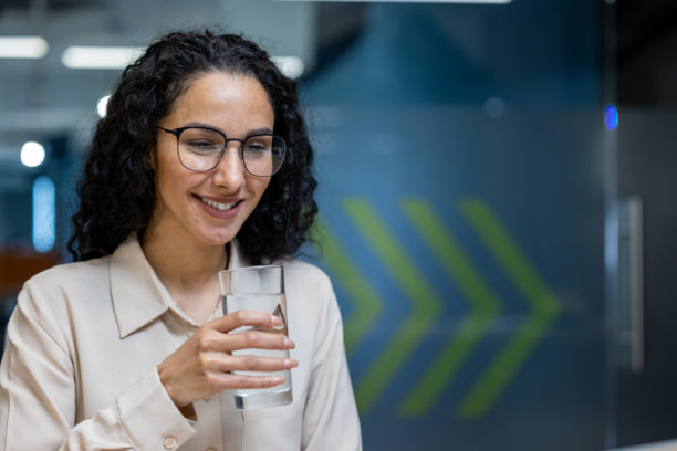 微笑的西班牙裔女商人在办公室桌前享用一杯清爽的水，倡导工作场所的水分补充和健康。图片下载