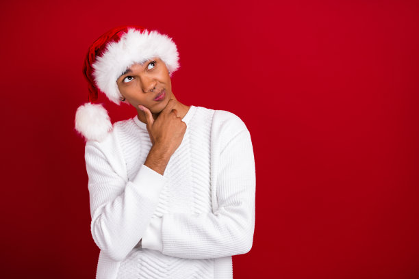年轻有趣的圣诞老人学生，撅着嘴，深思熟虑地看着空旷的空间，选择礼物，背景为红色。图片下载